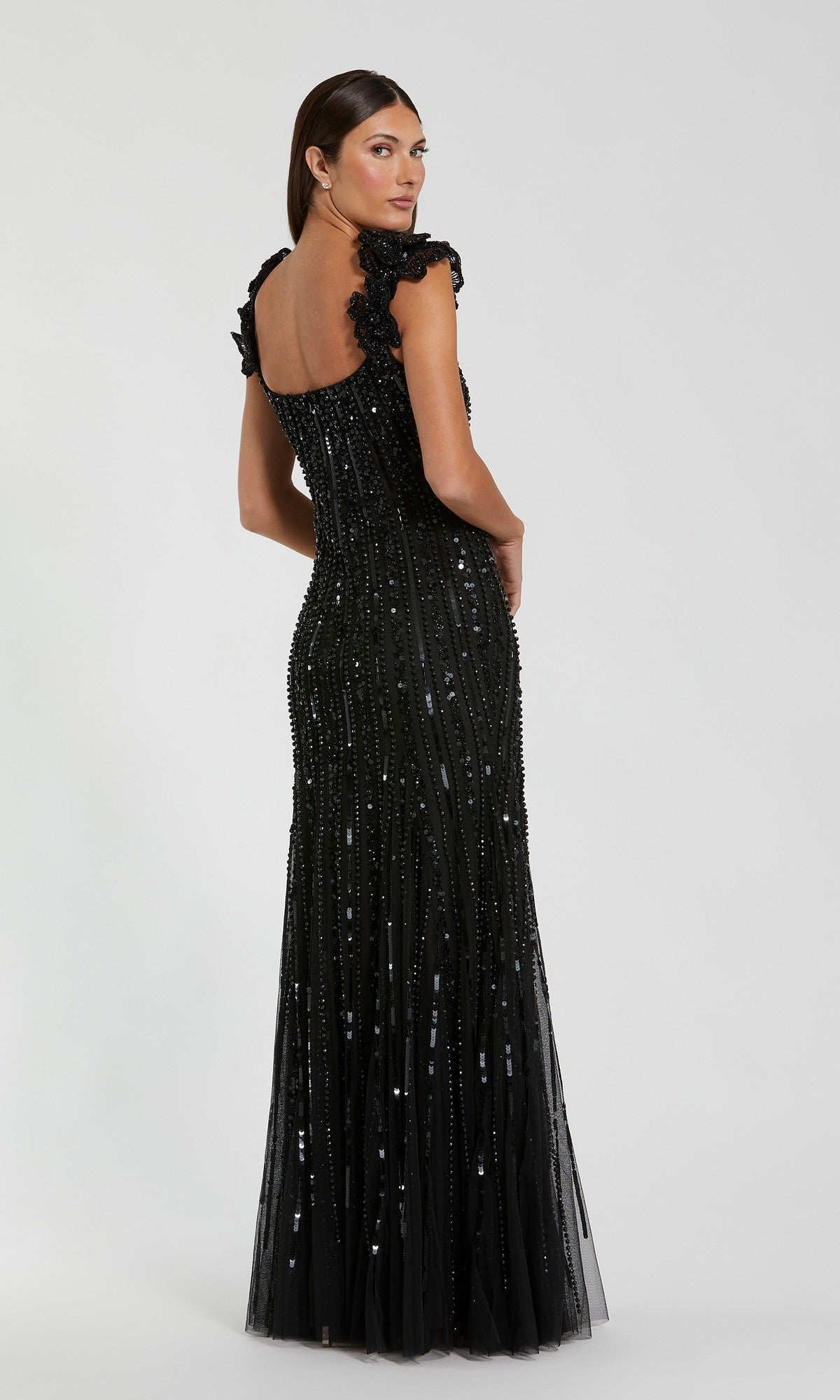 A woman wearing the Mac Duggal 94223 long formal dress—a floor-length, fitted black mesh gown adorned with beaded embellishments—stands with her back turned and looks over her shoulder against a plain background.